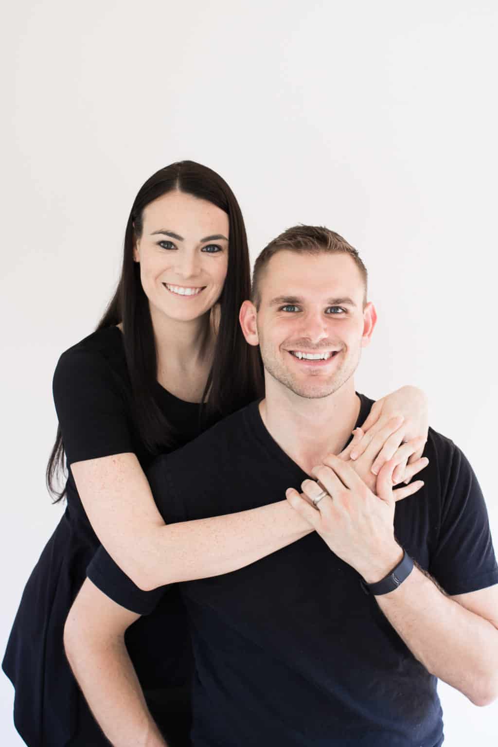 A smiling couple in matching black outfits, with the woman standing behind the man and her hands on his shoulders, posing for a happy portrait against a white backdrop.