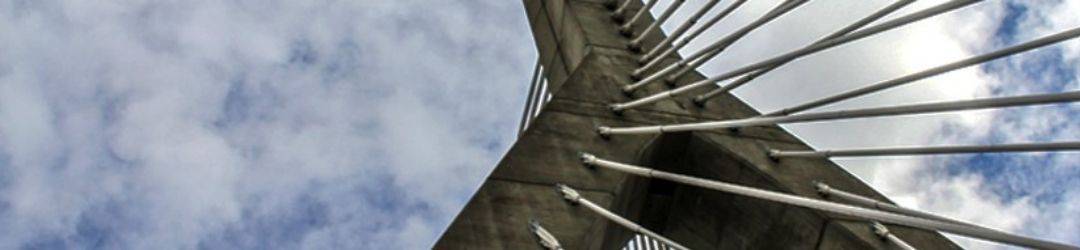 The photo shows a worm's-eye view of a modern cable-stayed bridge spanning the sky, with its towering pylon and radiating cables against a backdrop of scattered clouds.