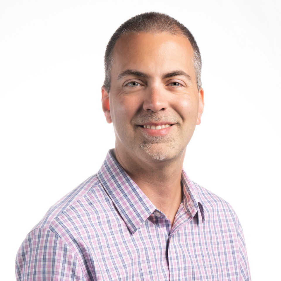 A confident man with a friendly smile wearing a plaid shirt.