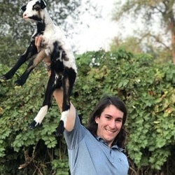 A smiling person proudly lifting a baby goat into the air outdoors, with a leafy hedge in the background.