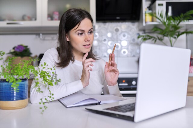 A focused woman holding a pen while engaging in a video call on her laptop in a bright kitchen setting.