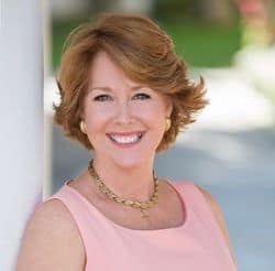 A smiling woman with shoulder-length hair, sporting a pale pink top and a gold necklace, against a blurred background.