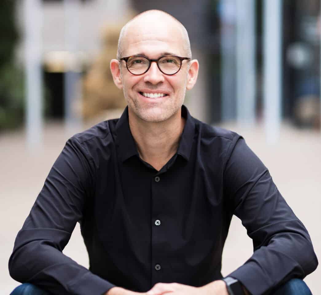A cheerful man with glasses wearing a black shirt smiles at the camera, exuding confidence and approachability.