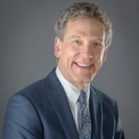A confident professional man with a warm smile, clad in a business suit and tie, posing against a grey background.