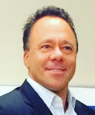 A smiling man with short, curly hair, dressed in a business casual style with a blazer over a collared shirt.
