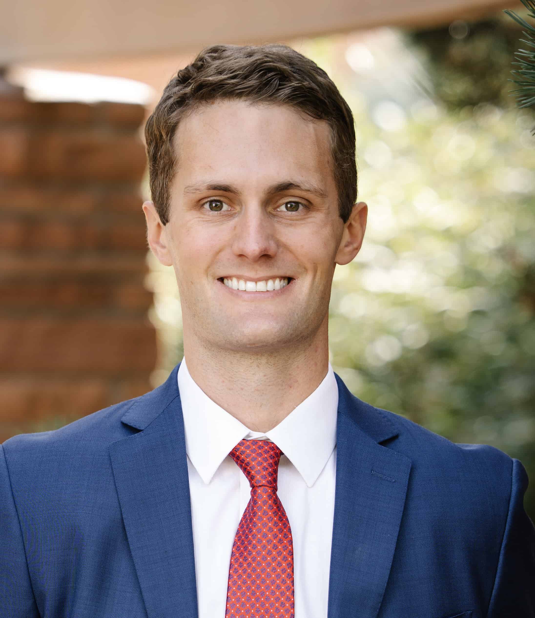 A professionally dressed man with a friendly smile, wearing a blue suit with a red tie, standing against a blurred natural backdrop.