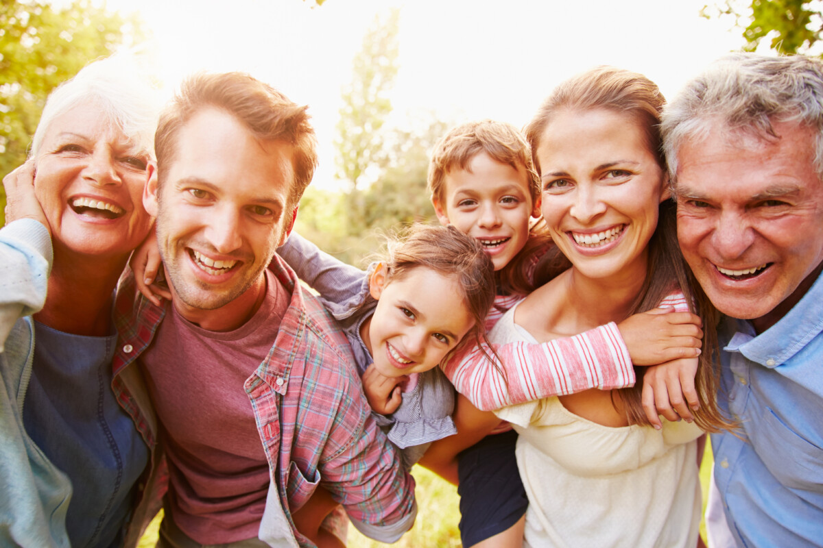 A joyful multi-generational family enjoying a sunny day outdoors, with grandparents, parents, and children smiling and hugging in a close-knit group.