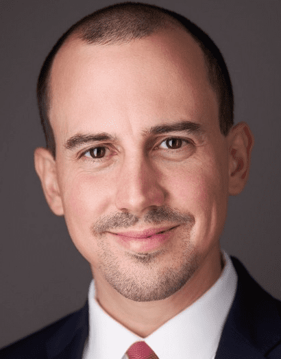 A professional headshot of a man with a friendly smile, dressed in business attire with a dark suit and a red tie.