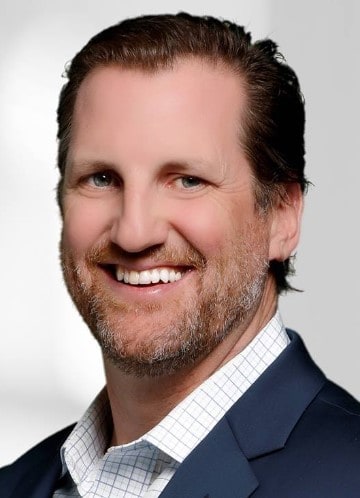 Professional man with a friendly smile wearing a dark suit and checkered shirt against a gray background.