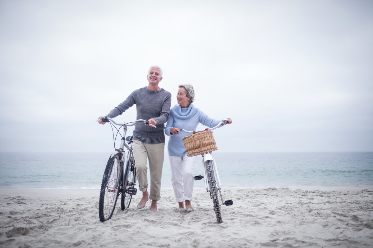 A cheerful senior couple walking with their bicycles on a sandy beach, enjoying a cloudy day by the shore.