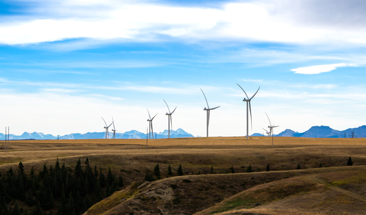 Wind turbines stand tall on a rolling grassland against a backdrop of distant mountain peaks, harnessing the power of nature beneath a spacious sky.