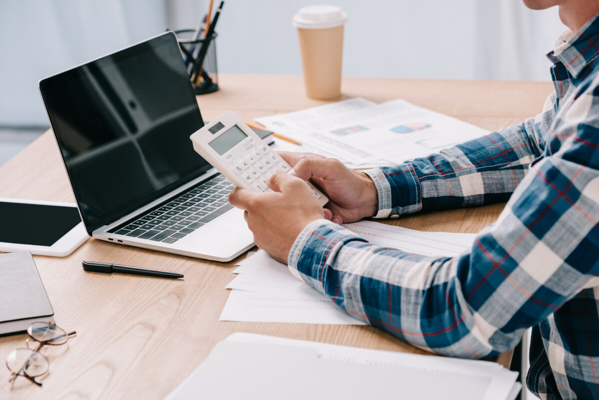 A person in a plaid shirt working on finances with a calculator, surrounded by paperwork and a laptop on a wooden desk.
