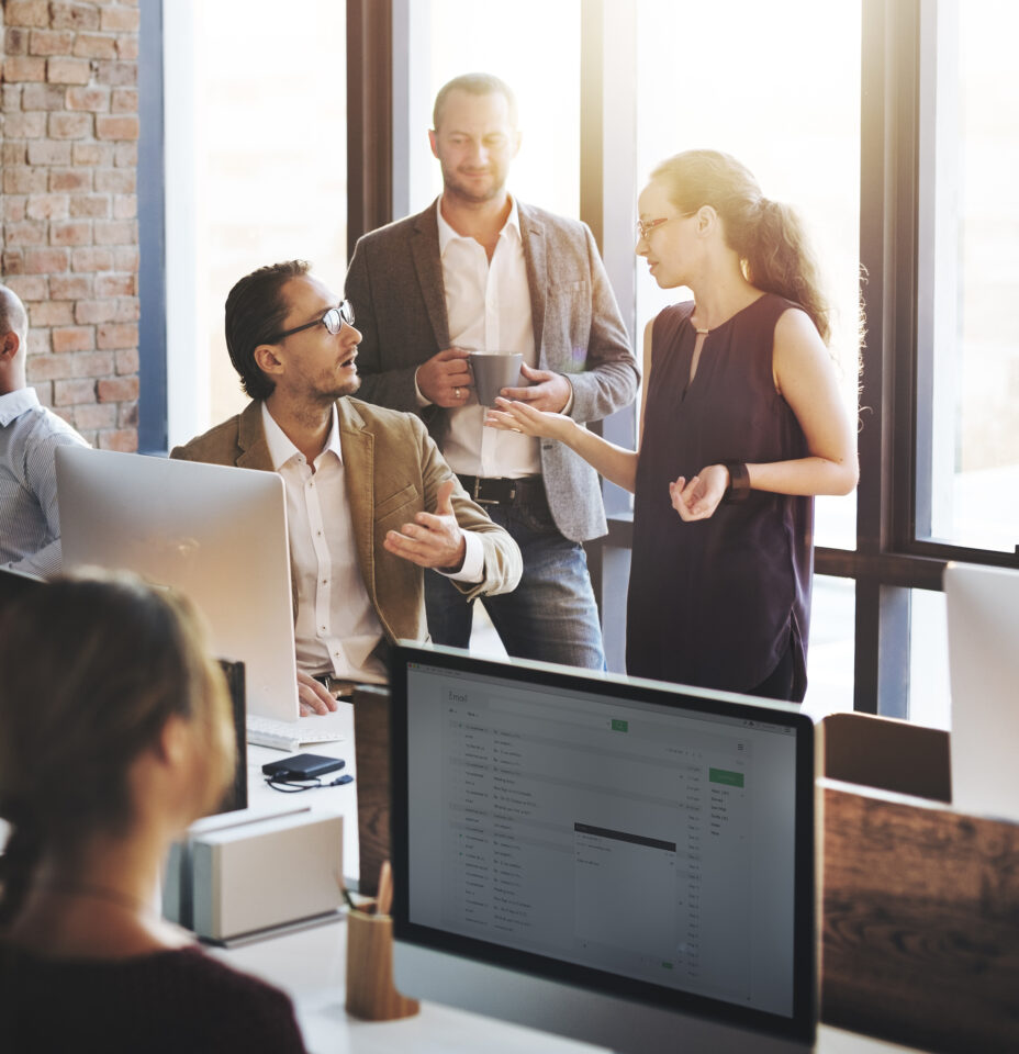 A group of professionals engaged in a lively discussion at a modern office space, with computers and casual attire suggesting a collaborative work environment.