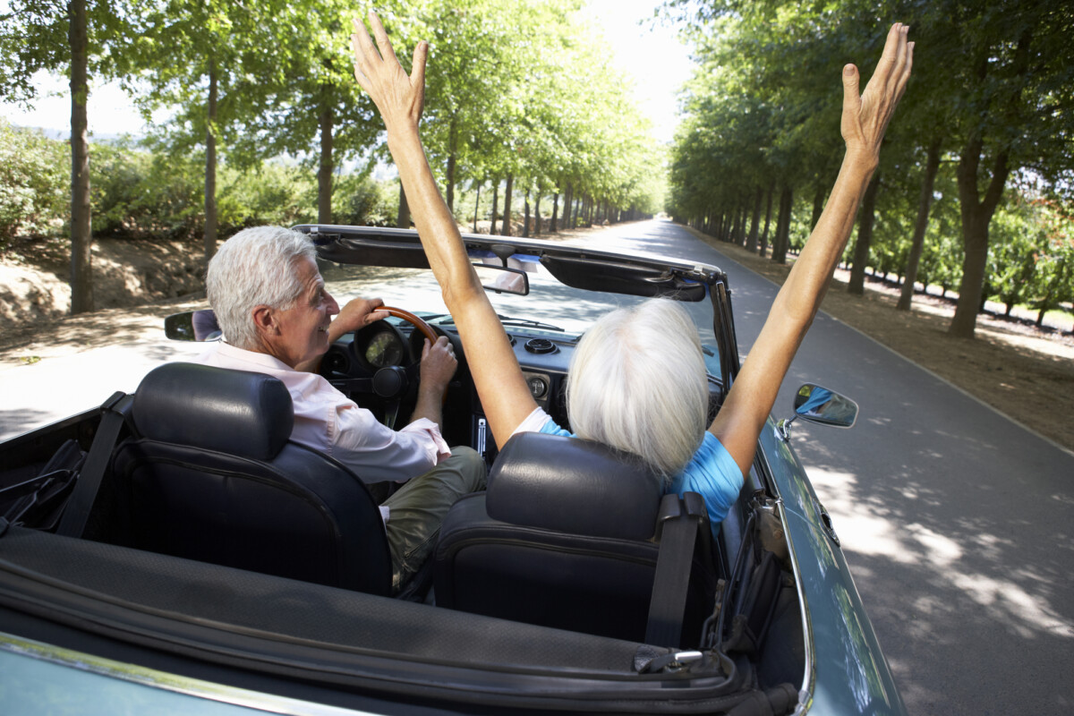 Senior couple enjoying a carefree ride in a convertible on a sunny day, with the woman joyfully raising her arms in the air.