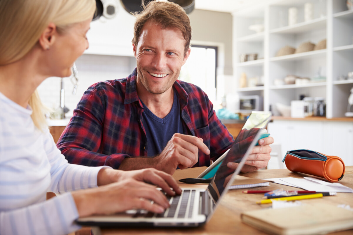 Two individuals collaborating at a home office with a friendly atmosphere; one uses a laptop while the other holds a tablet, both engaged in a productive conversation.