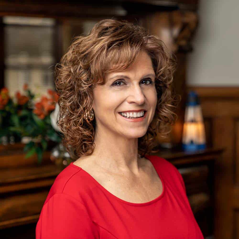 A smiling woman with curly hair, wearing a red top, against the backdrop of an interior room with wooden furniture and flowers in the background.