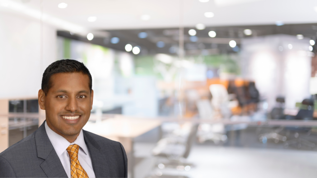 A confident professional man smiling in the foreground, with a modern, blurred office space in the background.