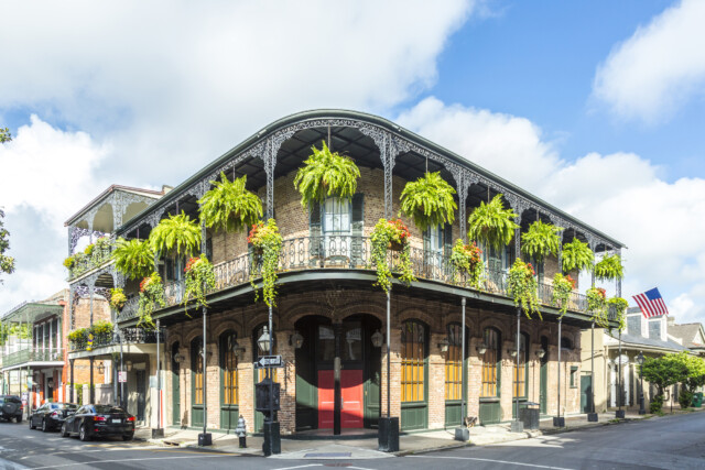 A charming two-story corner building in the french quarter with intricate wrought-iron balconies adorned with lush green plants and an american flag fluttering in the breeze.
