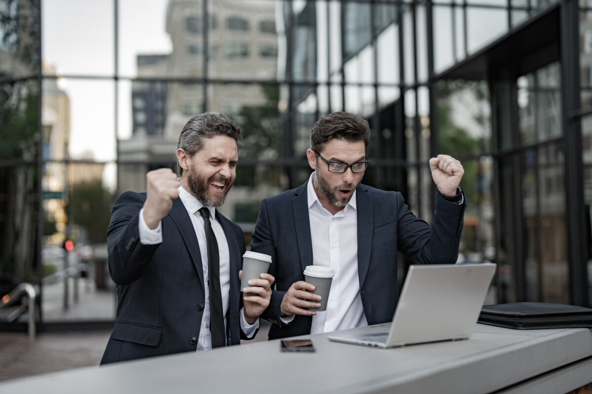 Victorious moment: two professional men in suits celebrating success with coffee in hand, in front of a laptop outdoors, against an urban backdrop.