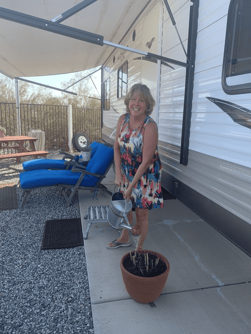 A person smiling while standing outside an rv, watering a plant in a pot with a watering can.
