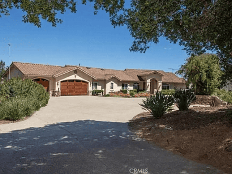 Spacious single-story home with an expansive driveway and attached garage, set against a backdrop of lush greenery under a clear blue sky.