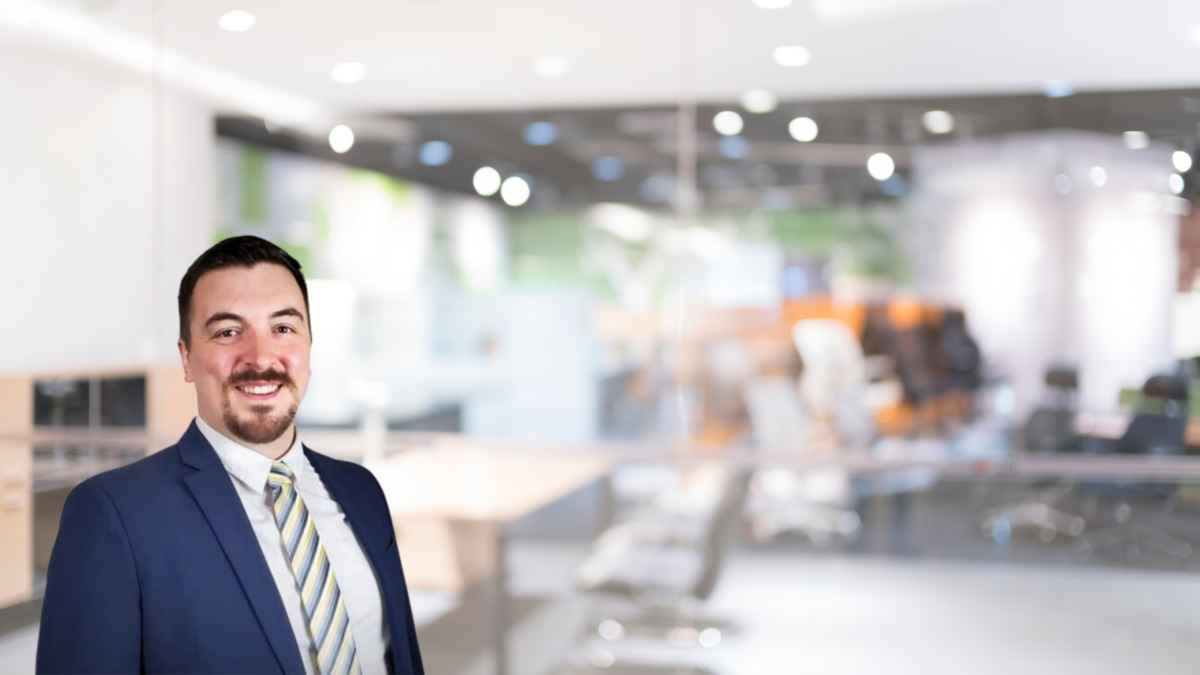 A smiling man in a blue suit and striped tie stands in a modern, brightly lit office space with blurred background. The office features contemporary furnishings, including desks and chairs, and has a clean, professional atmosphere.
