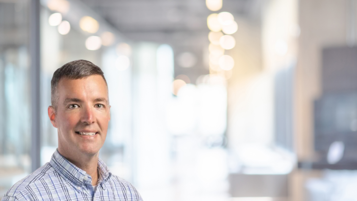 A man wearing a blue checkered shirt stands smiling in a modern, warmly lit office setting. Blurred background features desks, chairs, and hanging lights, indicating a spacious, open workspace.