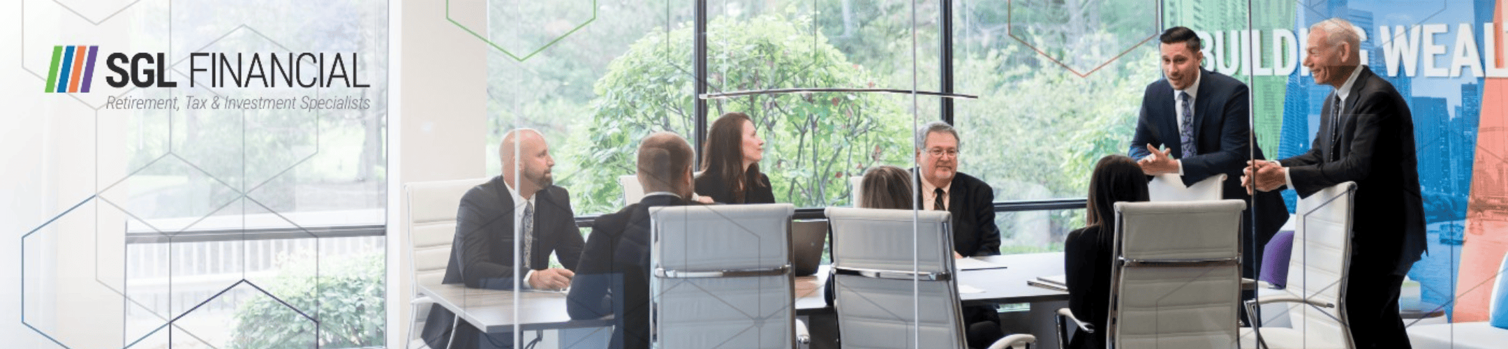A group of people are seated around a conference table in a modern office setting. Two individuals are standing and speaking to the group. The office has large windows with a view of greenery outside. The text 