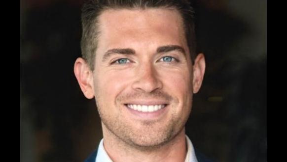 A man with short brown hair and blue eyes is smiling at the camera. He is wearing a dark blue blazer and a white shirt, and the background is blurred and dark.