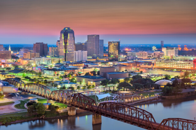 A vibrant nighttime view of Shreveport, Louisiana, showcasing a well-lit skyline with buildings of varying heights. The colorful lights reflect on the river below, and a historic railway bridge spans the water in the foreground. The sky has a warm, gradient sunset.