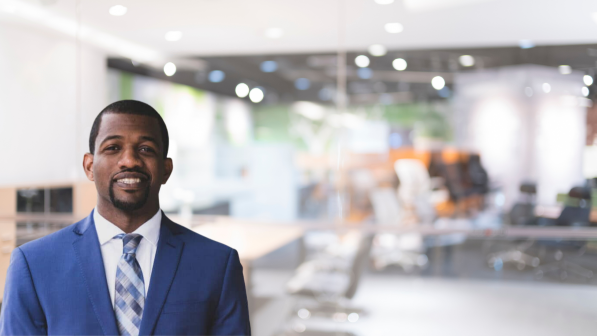 A man in a blue suit and striped tie smiles while standing in a modern, brightly lit office with glass walls and blurred background.