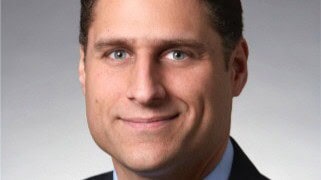 A man in a suit and tie with short brown hair is smiling slightly at the camera. The background is plain and light-colored.