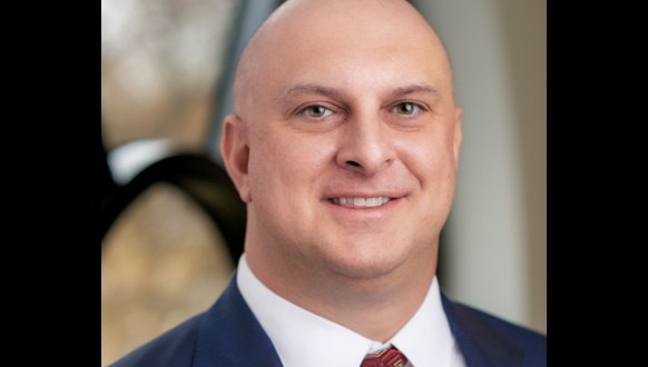 A man with a shaved head in a blue suit, white shirt, and patterned red tie, smiling at the camera. The background is softly blurred with neutral tones and a window.