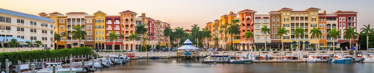 Colorful waterfront buildings with palm trees line a marina filled with boats, under a clear sky at sunset. A blue-and-white gazebo sits at the center near the water’s edge.