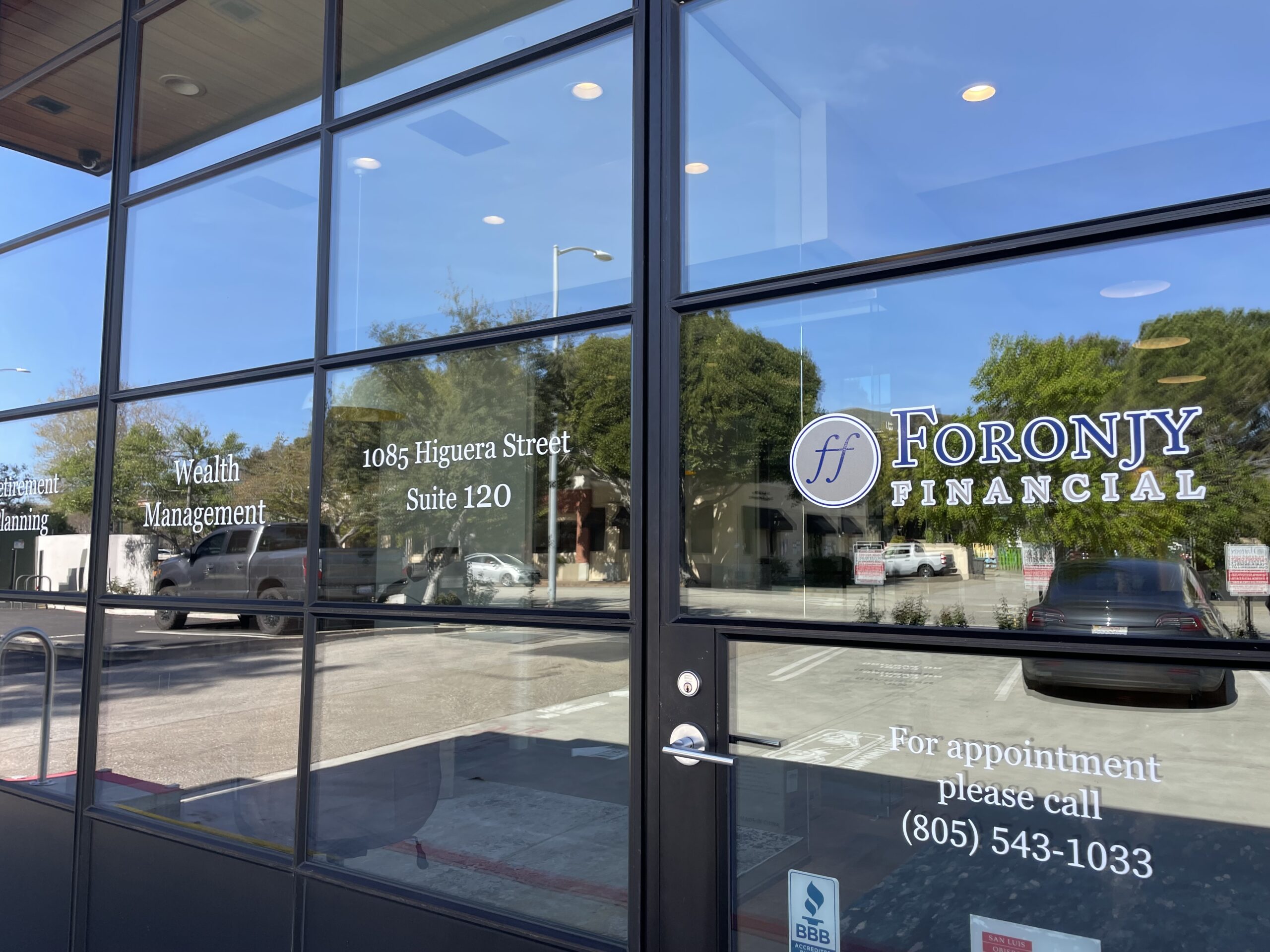 The glass front of a financial office, showing the Foronjy Financial logo, address (1085 Higuera Street, Suite 120), and contact number (805-543-1033). Reflections of cars and trees are visible in the glass.