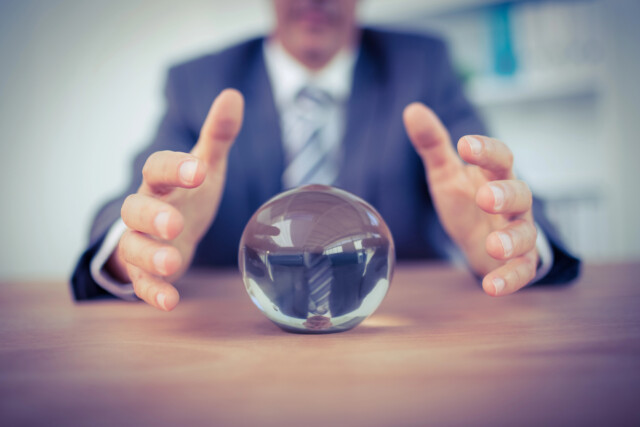 A person in a suit sits at a desk with hands hovering around a clear glass crystal ball, suggesting prediction or foresight. The focus is on the crystal ball and hands, with the person's face blurred.