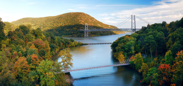 A scenic view of a river winding through a forest with trees in autumn colors, two bridges crossing the river, and a green, tree-covered hill in the background under a partly cloudy sky.