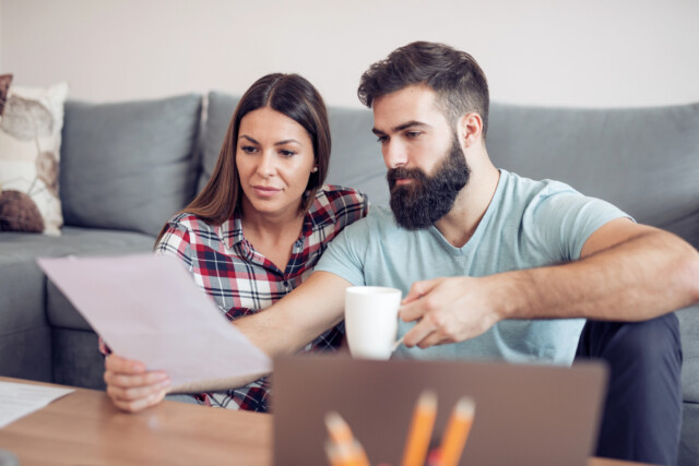 A couple sits on the floor in front of a coffee table, looking at a document together. The man holds a white mug, and a laptop with pencils in a holder is visible in the foreground.