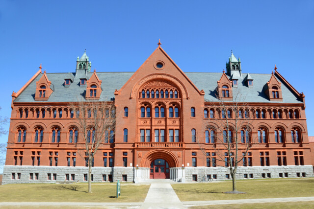 A large, symmetrical red brick building with arched windows and detailed stonework, featuring multiple peaked roofs and small towers, set against a clear blue sky with trees and a walkway in front.