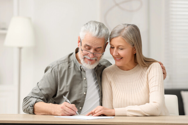 An older man and woman sit closely together at a table, smiling and looking at papers as the man writes. The woman has her arm around the man in a bright, cozy room.
