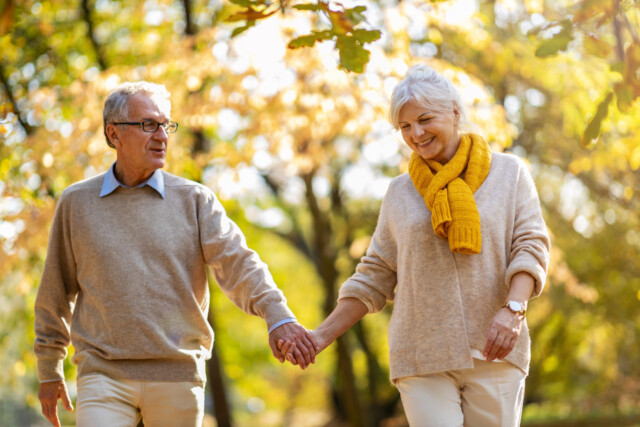 An older couple walks hand in hand outdoors on a sunny fall day. They are smiling and wearing light sweaters; the woman has a yellow scarf. Autumn trees with golden leaves are in the background.