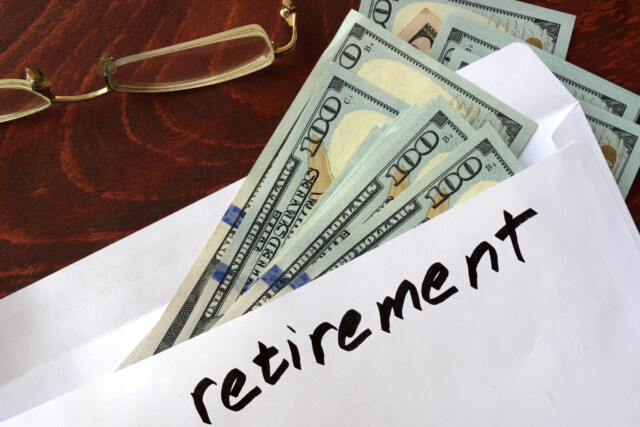 An envelope labeled "retirement" filled with several $100 bills sits on a wooden surface next to a pair of eyeglasses.