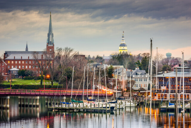 A marina with docked sailboats sits in front of a townscape featuring historic buildings, a church with a tall steeple, and a domed capitol building under a cloudy sky.