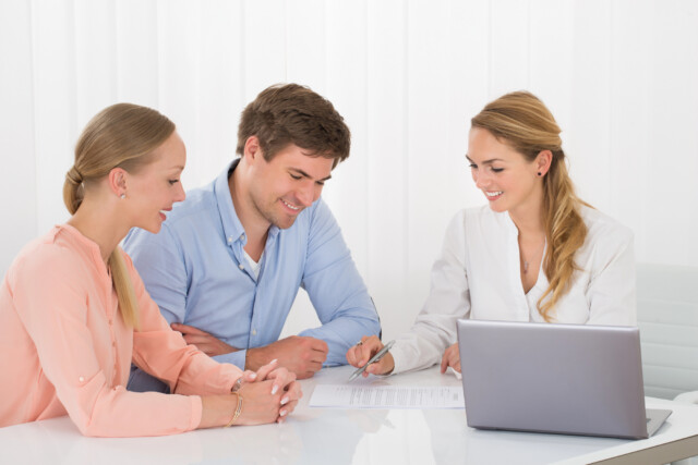 A smiling couple sits at a desk with a professional woman who is showing them a document to sign. A laptop is open on the desk. All three are engaged in a friendly discussion in a bright office setting.