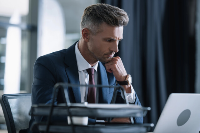 A man in a suit sits at a desk, resting his chin on his hand and looking intently at a laptop screen, appearing focused and deep in thought. Office folders are visible in the foreground.