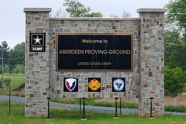 A stone sign reads "Welcome to ABERDEEN PROVING GROUND UNITED STATES ARMY" with four U.S. Army logos below, set outdoors with grass and trees in the background.