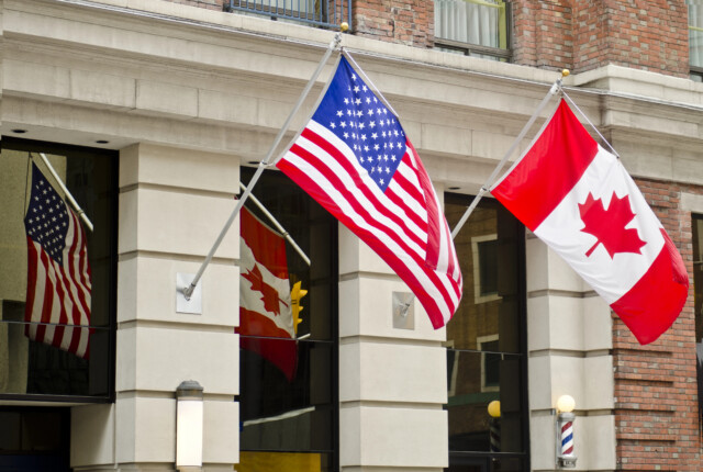 Two flags, the United States flag and the Canadian flag, are mounted on the exterior of a brick building. Their reflections are visible in the building's large windows.